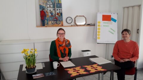 Sarah Klein und Christian Schega sitzen amTisch, vor sich weißes Papier und orange Papierzuschnitte, außerdem eine Schale mit Osterglocken. Im Hintergrund ein Bild, gemalt von Doris Zorn, eine Uhr und ein Flipchart mit mehren Zetteln und Moderationskarten.